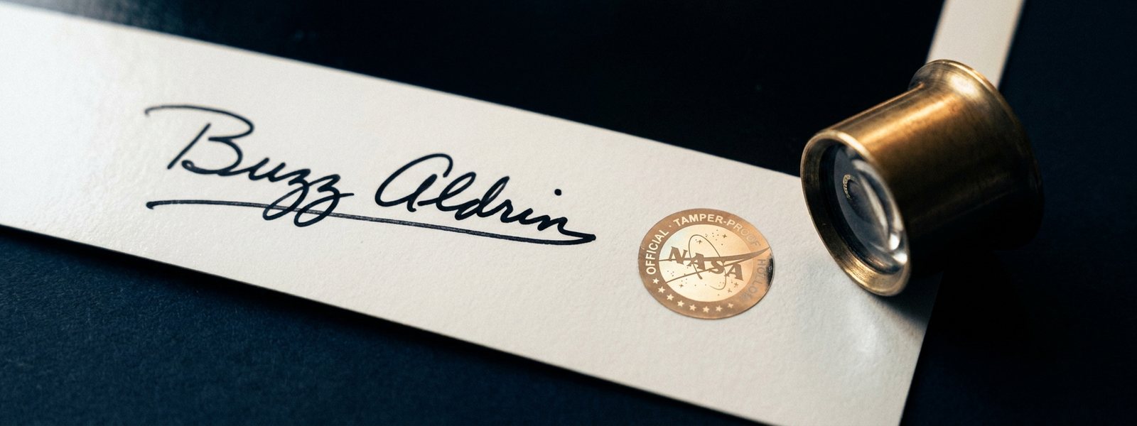 Macro of a vintage space photograph margin showing a handwritten astronaut signature and a round tamper-proof authentication hologram beside a brass loupe