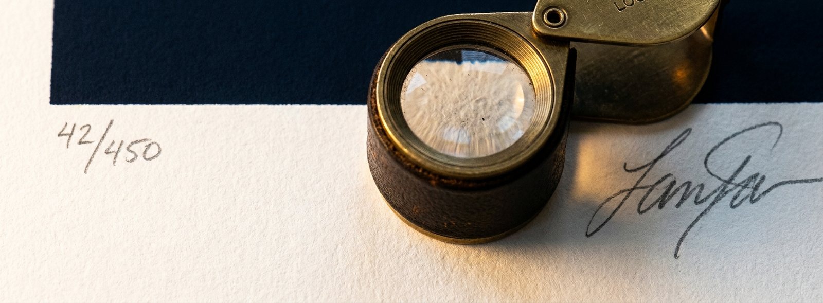 Macro of a print margin: a pencil edition number, a signature and a brass loupe on cotton paper
