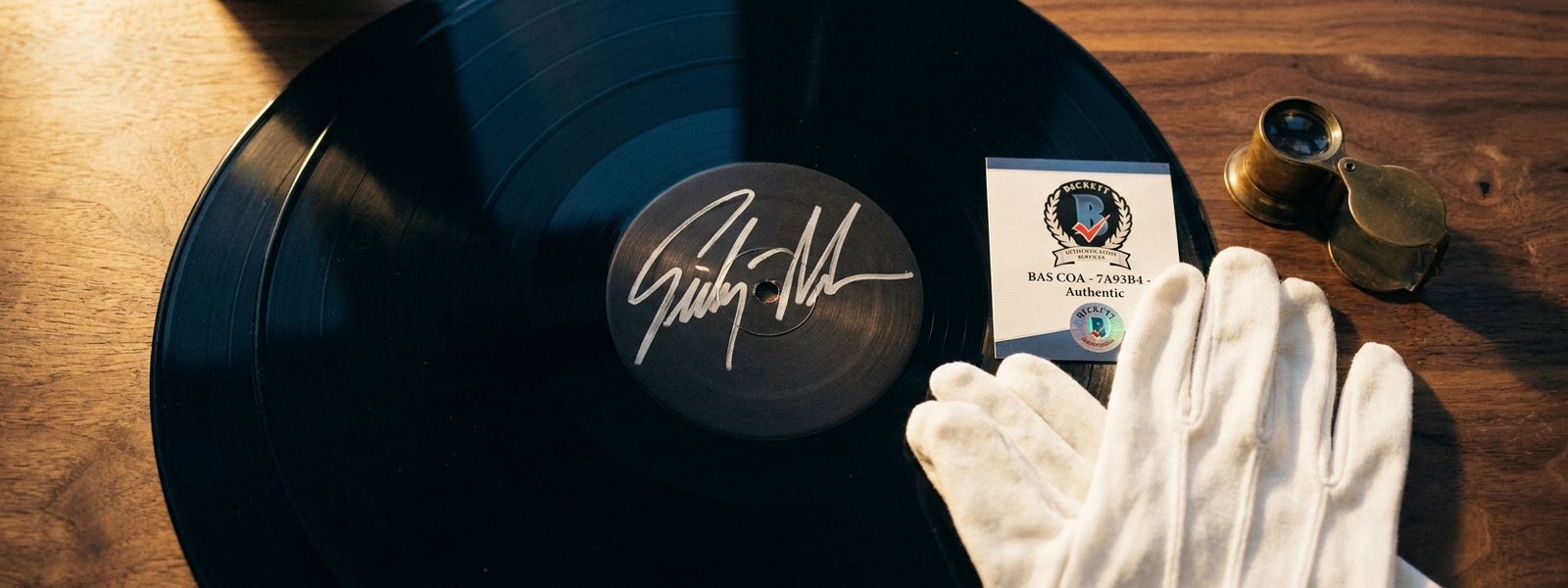 A signed vinyl record with a silver-ink signature beside a Beckett-style authentication card, white cotton gloves, and a brass loupe